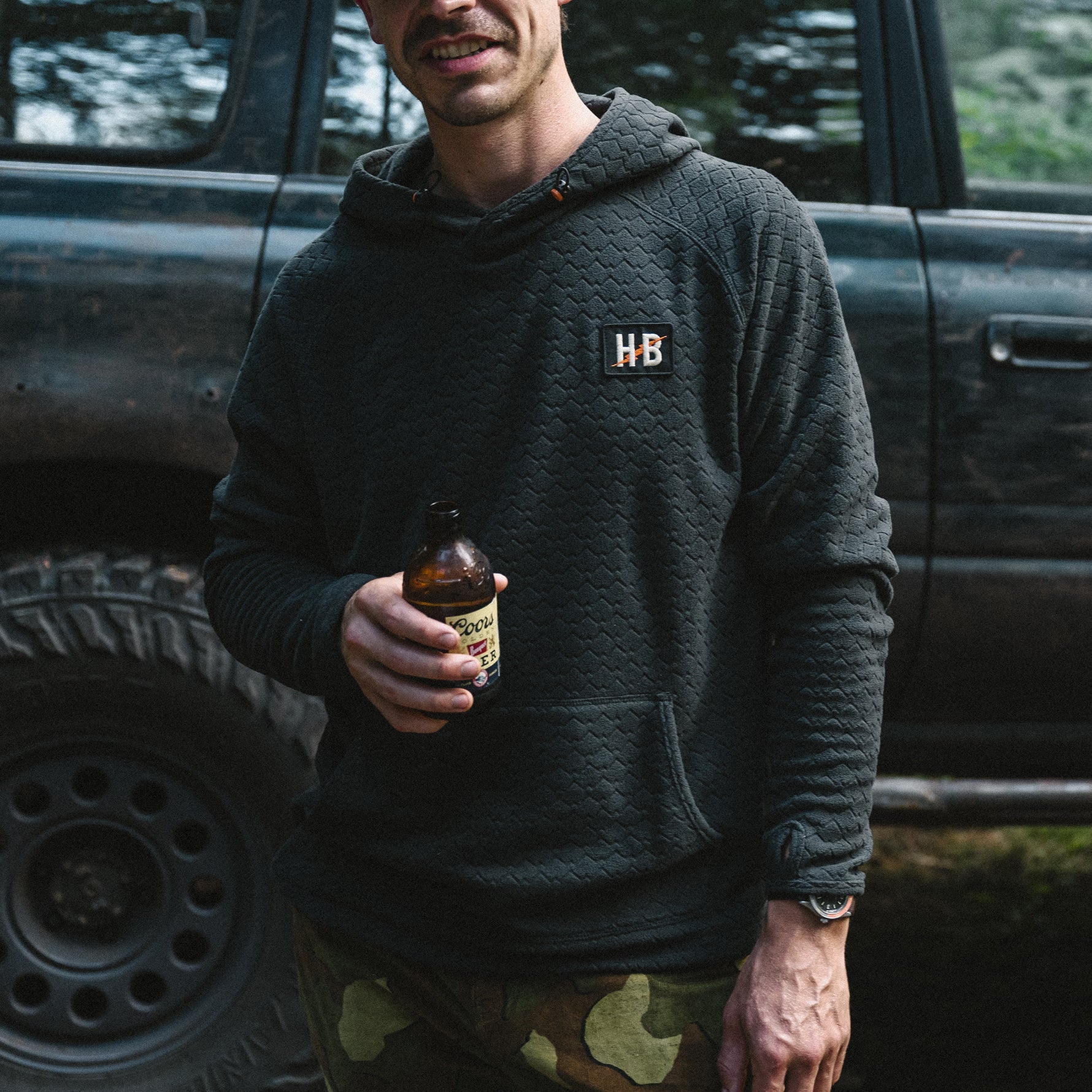Man holding a bottle in front of a truck with an 'HB' logo on his hoodie.