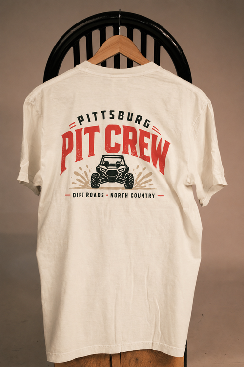 An off-white short-sleeve t-shirt hanging on a wooden hanger against a black chair, featuring a large red and black graphic on the back that reads 'PITTSBURG PIT CREW' with an off-road vehicle illustration.