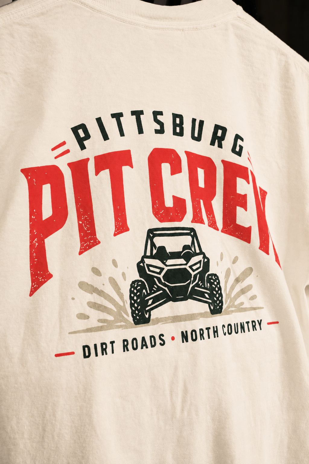 Close-up of a white graphic t-shirt featuring a red and black 'PIT CREW' print with an off-road vehicle illustration and 'DIRT ROADS NORTH COUNTRY' text.