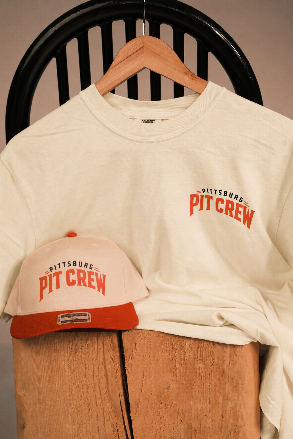 An off-white crewneck t-shirt with a red and black 'Pittsburgh Pit Crew' graphic on the left chest, displayed on a wooden hanger next to a matching off-white and red baseball cap on a wooden stand.