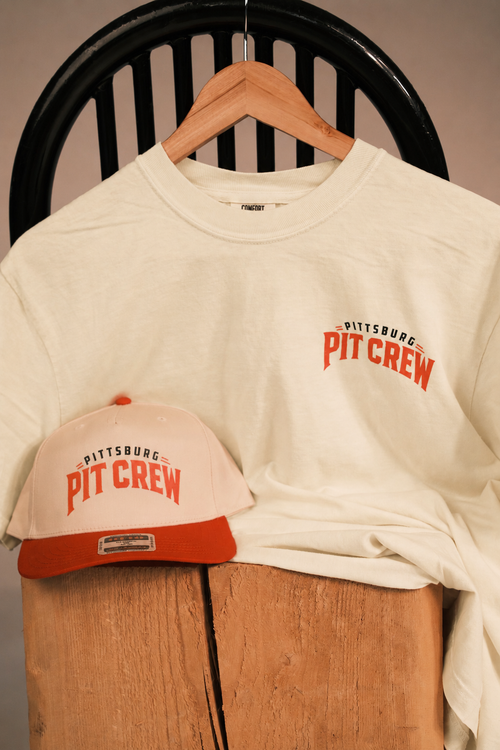 An off-white crewneck t-shirt with a red and black 'Pittsburgh Pit Crew' graphic on the left chest, displayed on a wooden hanger next to a matching off-white and red baseball cap on a wooden stand.