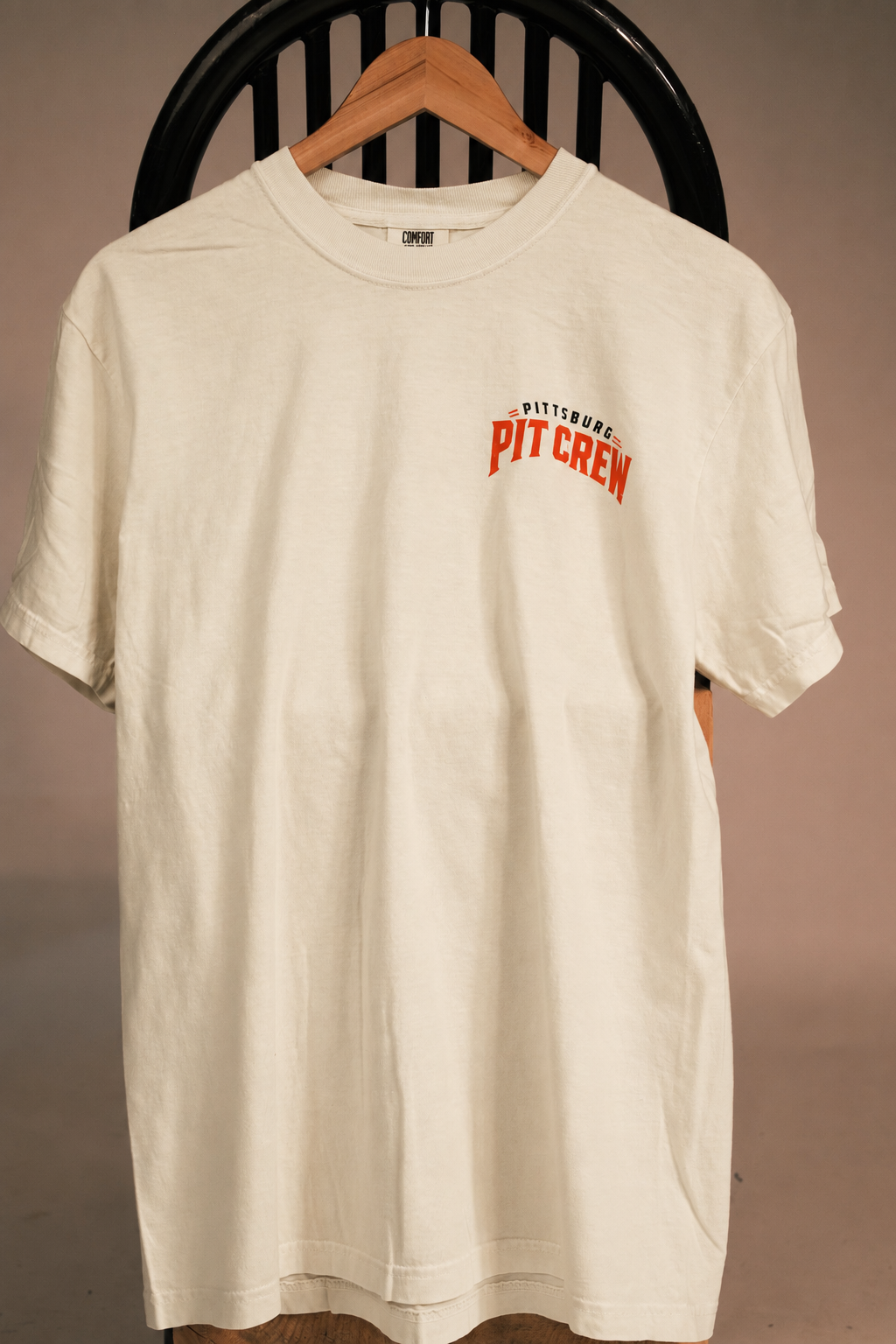 An off-white short-sleeve t-shirt hanging on a wooden hanger against a dark chair, featuring 'PIT CREW' and 'BOSTON' printed in red and black on the left chest.