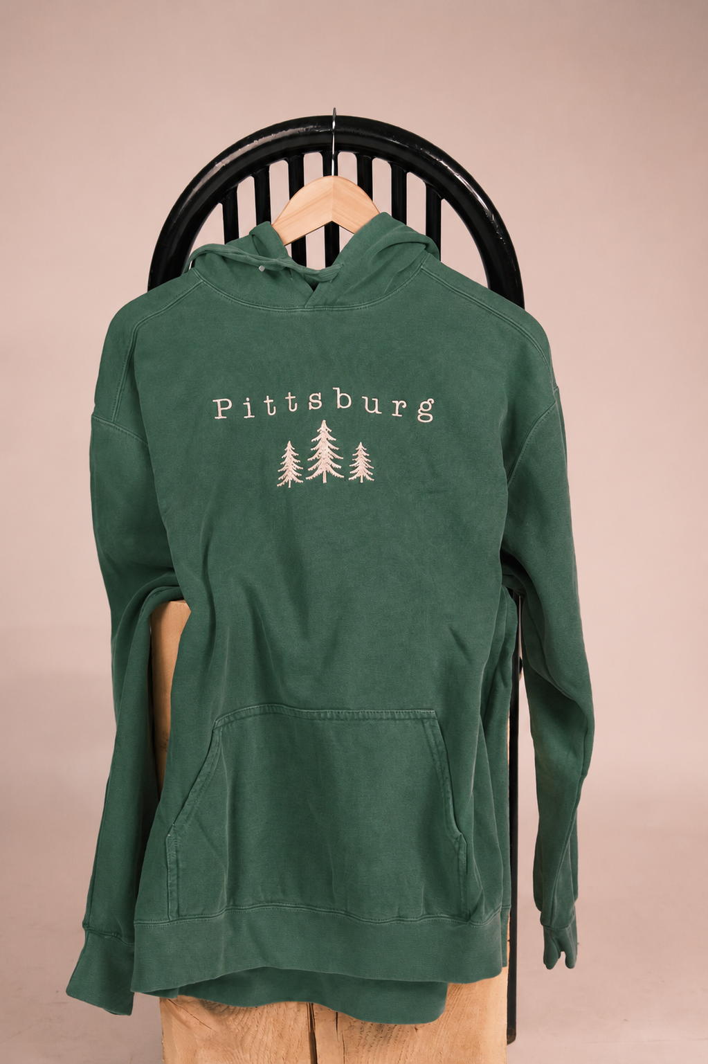 A dark green pullover hoodie with 'Pittsburg' and three pine tree graphics printed in white on the chest, hanging on a wooden hanger from a black chair against a solid beige background.