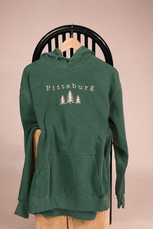A dark green pullover hoodie with 'Pittsburg' and three pine tree graphics printed in white on the chest, hanging on a wooden hanger from a black chair against a solid beige background.