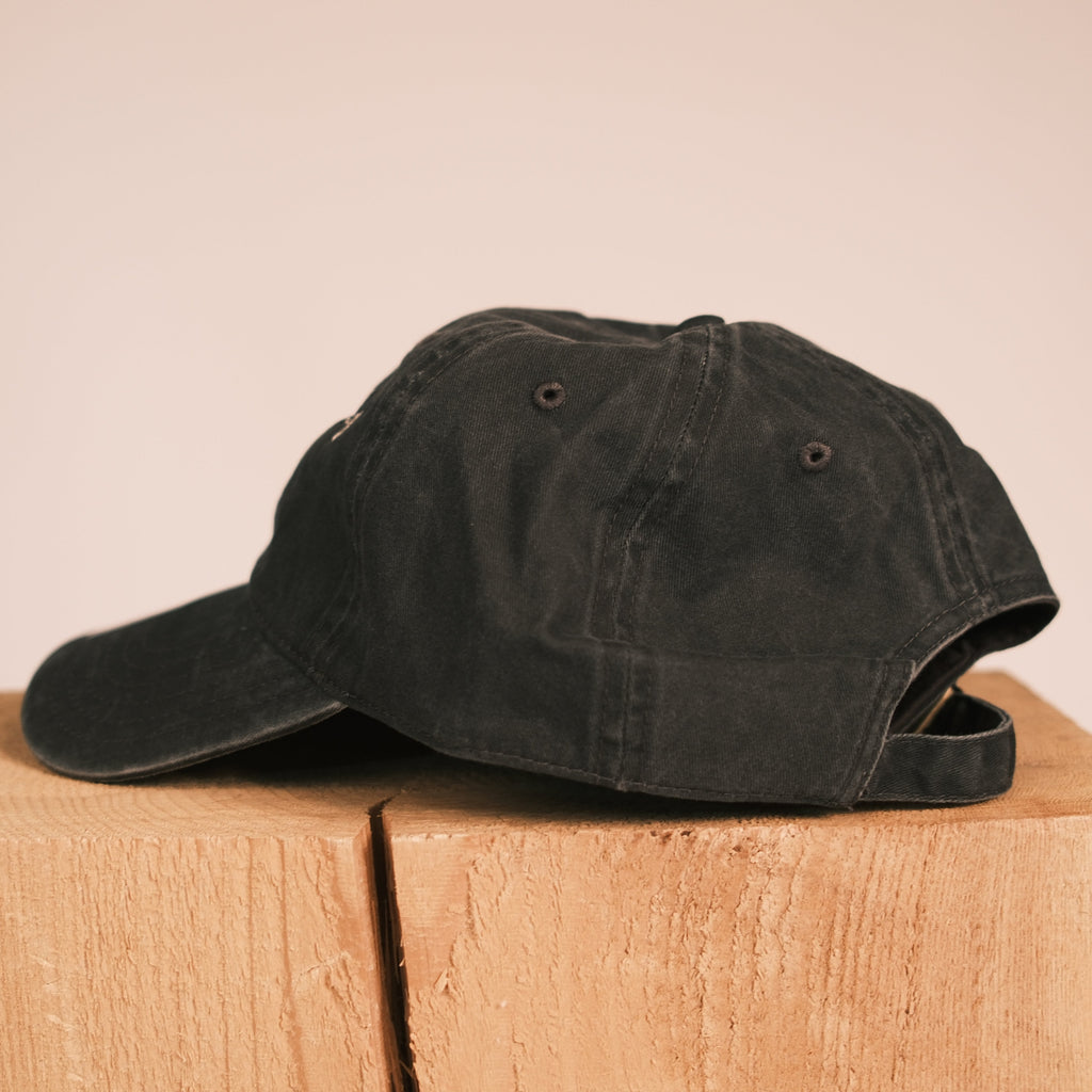 A black washed cotton baseball cap resting on a light wood block against a solid beige background.