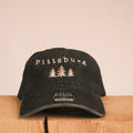 A black baseball cap with 'Pittsburg' and three pine trees embroidered on the front, displayed on a wooden block against a plain beige background.