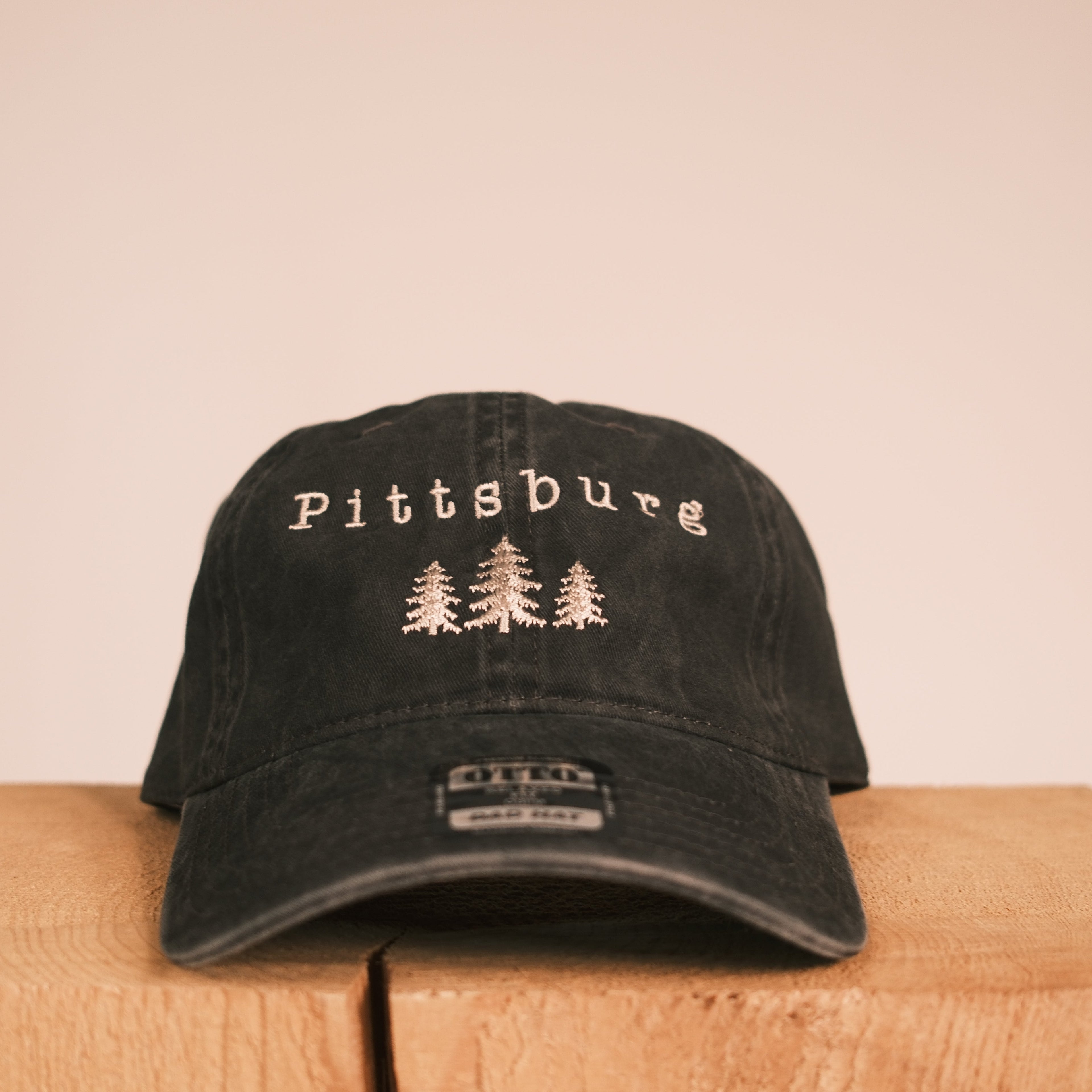 A black baseball cap with 'Pittsburg' and three pine trees embroidered on the front, displayed on a wooden block against a plain beige background.