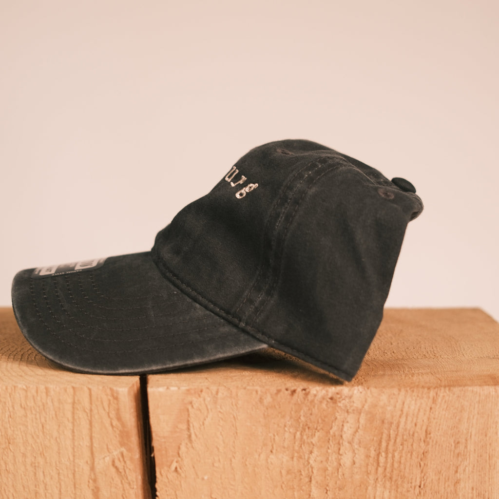 A black washed baseball cap with white embroidered lettering, resting on a light brown wooden block against a solid beige background.