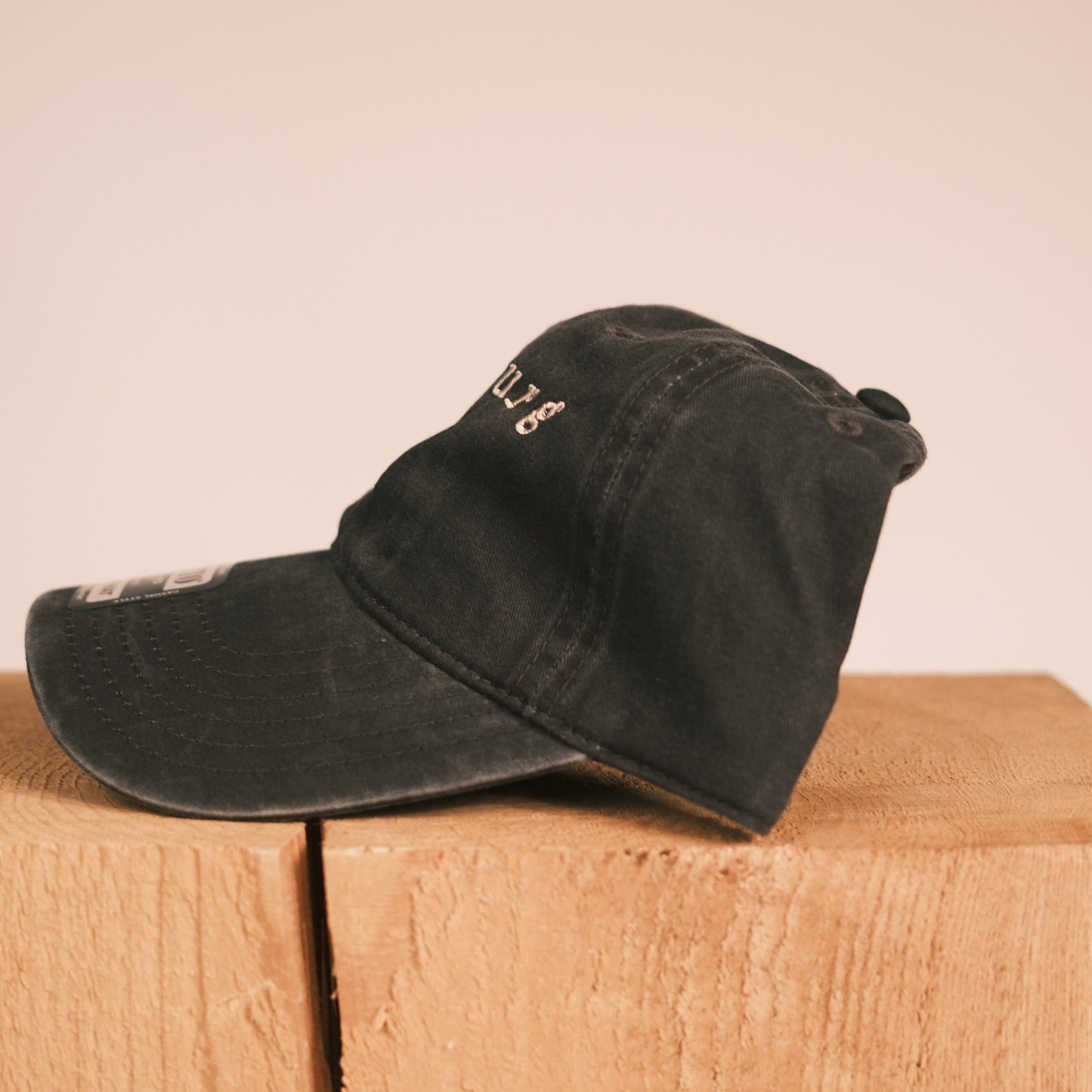 A black washed baseball cap with white embroidered lettering, resting on a light brown wooden block against a solid beige background.