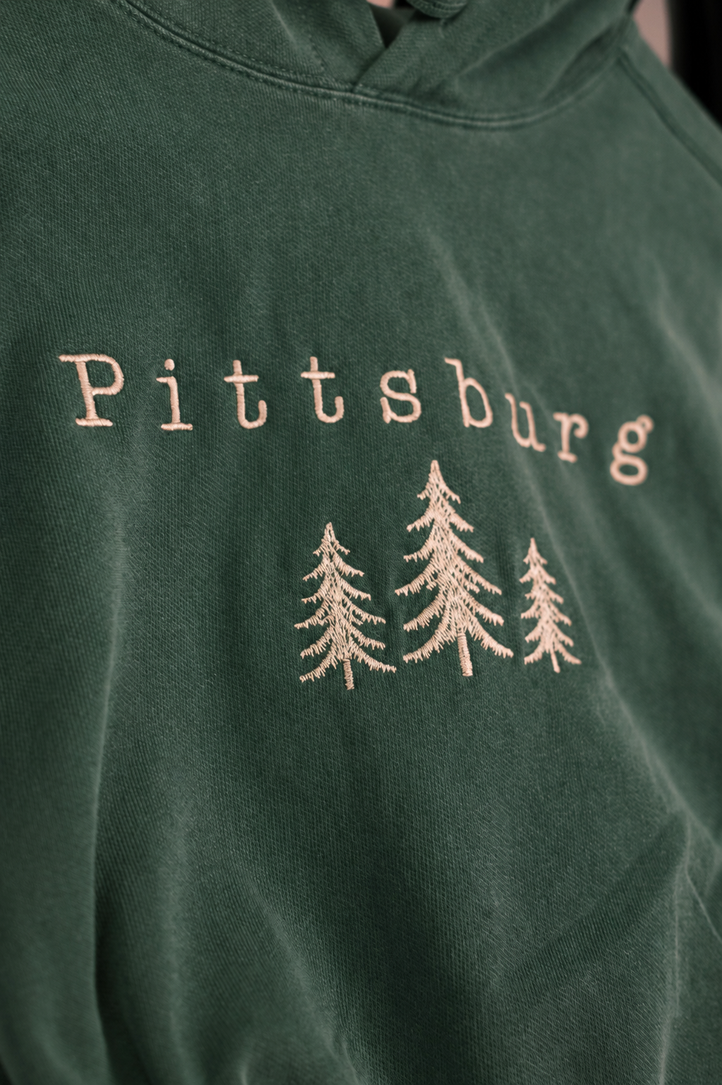 Close-up of a dark green hooded sweatshirt with 'Pittsburg' embroidered in a light beige tone above three matching pine tree silhouettes.