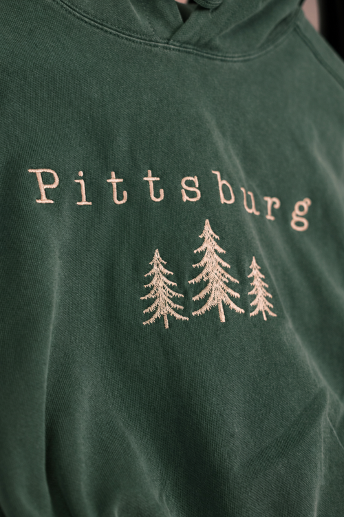 Close-up of a dark green hooded sweatshirt with 'Pittsburg' embroidered in a light beige tone above three matching pine tree silhouettes.
