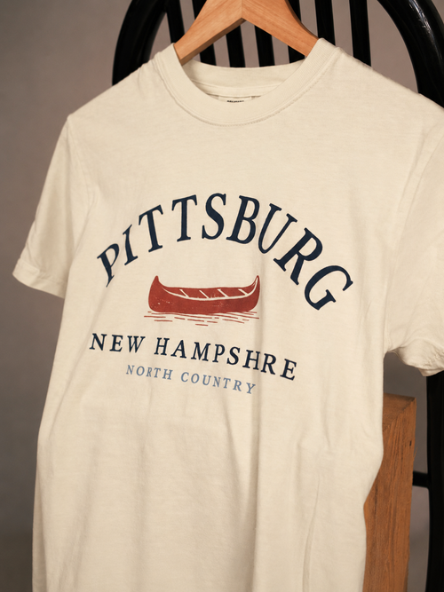 An off-white crewneck t-shirt hanging on a wooden hanger, featuring a graphic print with the words 'PITTSBURG NEW HAMPSHIRE NORTH COUNTRY' in navy blue and a red canoe illustration.