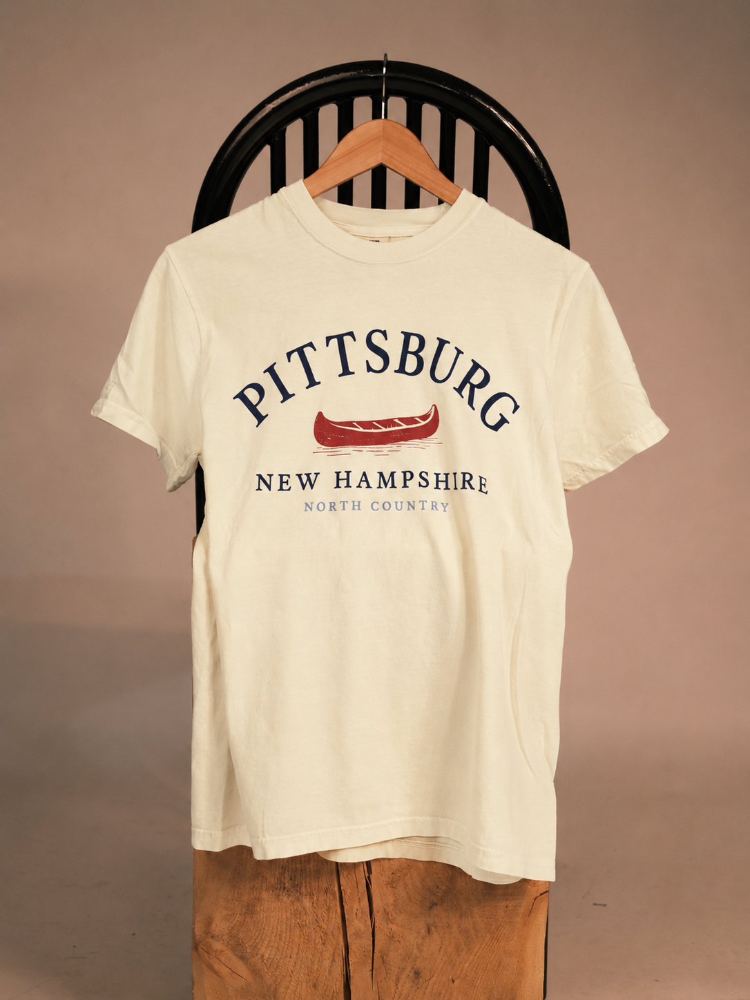 An off-white short-sleeve graphic t-shirt hanging on a wooden hanger against a dark wooden chair, featuring a red canoe graphic and the text 'PITTSBURG NEW HAMPSHIRE NORTH COUNTRY' in navy blue.