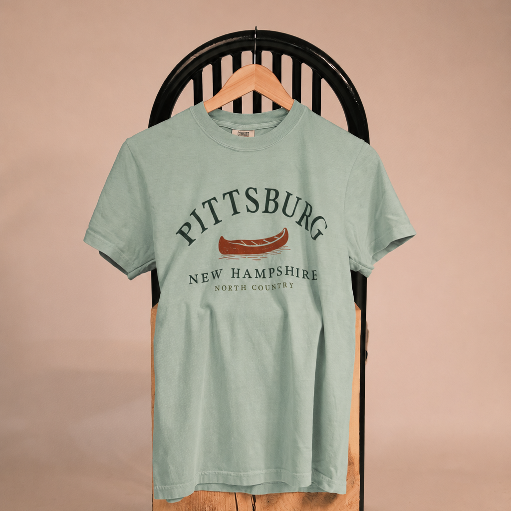 A light sage green graphic t-shirt hanging on a wooden hanger from a black slatted chair, displayed on a rustic wood block against a solid beige background. The shirt features a graphic print reading 'PITTSBURG NEW HAMPSHIRE' with a hot dog illustration.