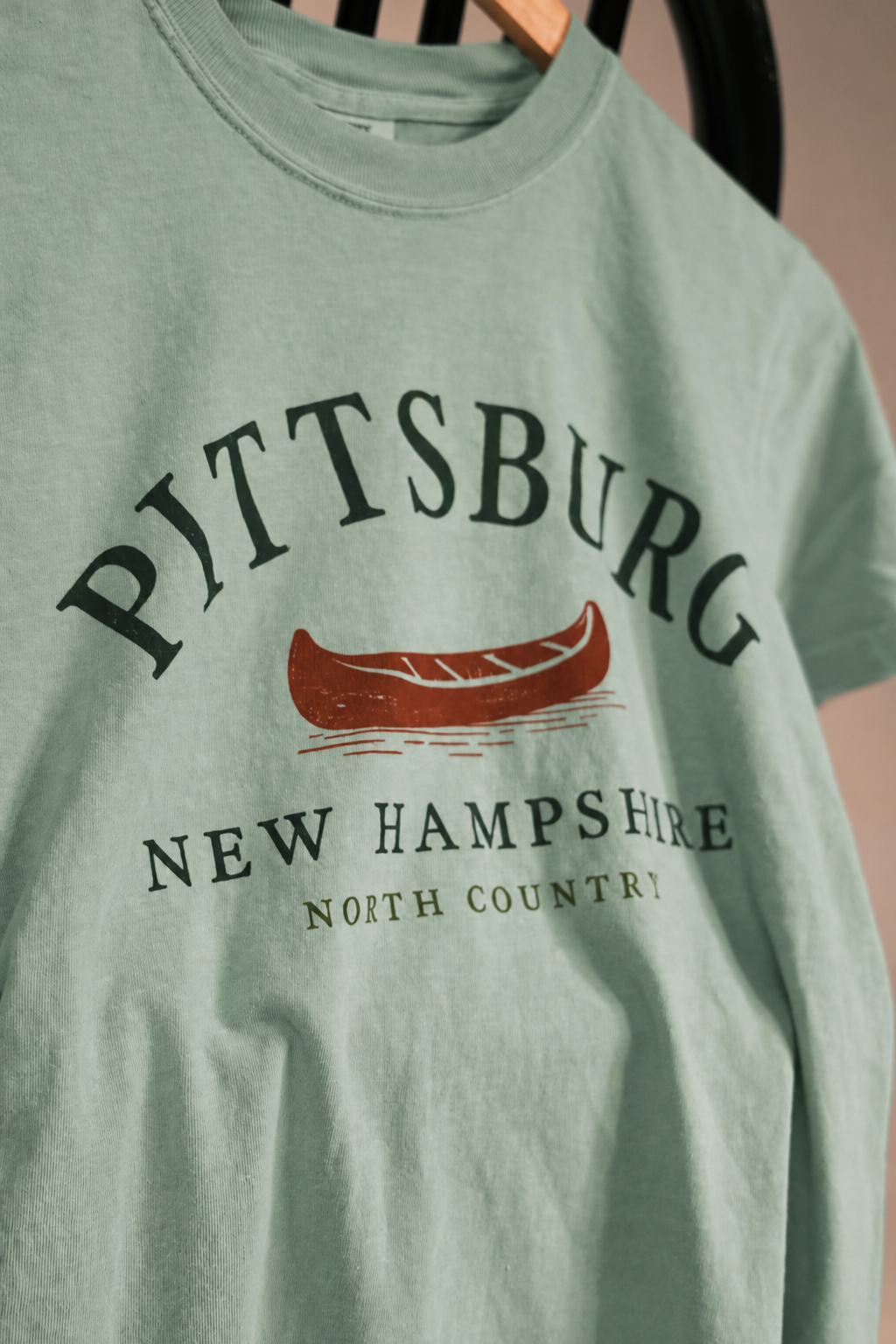 A close-up of a sage green t-shirt hanging on a wooden hanger, featuring a graphic print with the text 'PITTSBURG NEW HAMPSHIRE NORTH COUNTRY' and a red canoe illustration.