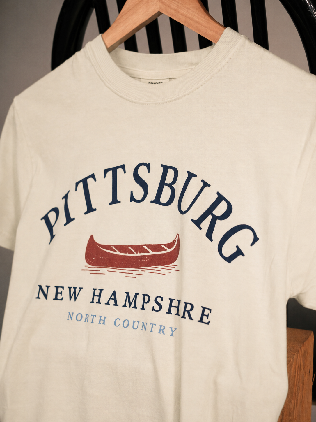 An off-white crewneck t-shirt hanging on a wooden hanger, featuring a graphic print with the text 'PITTSBURG NEW HAMPSHIRE NORTH COUNTRY' and a red canoe illustration.