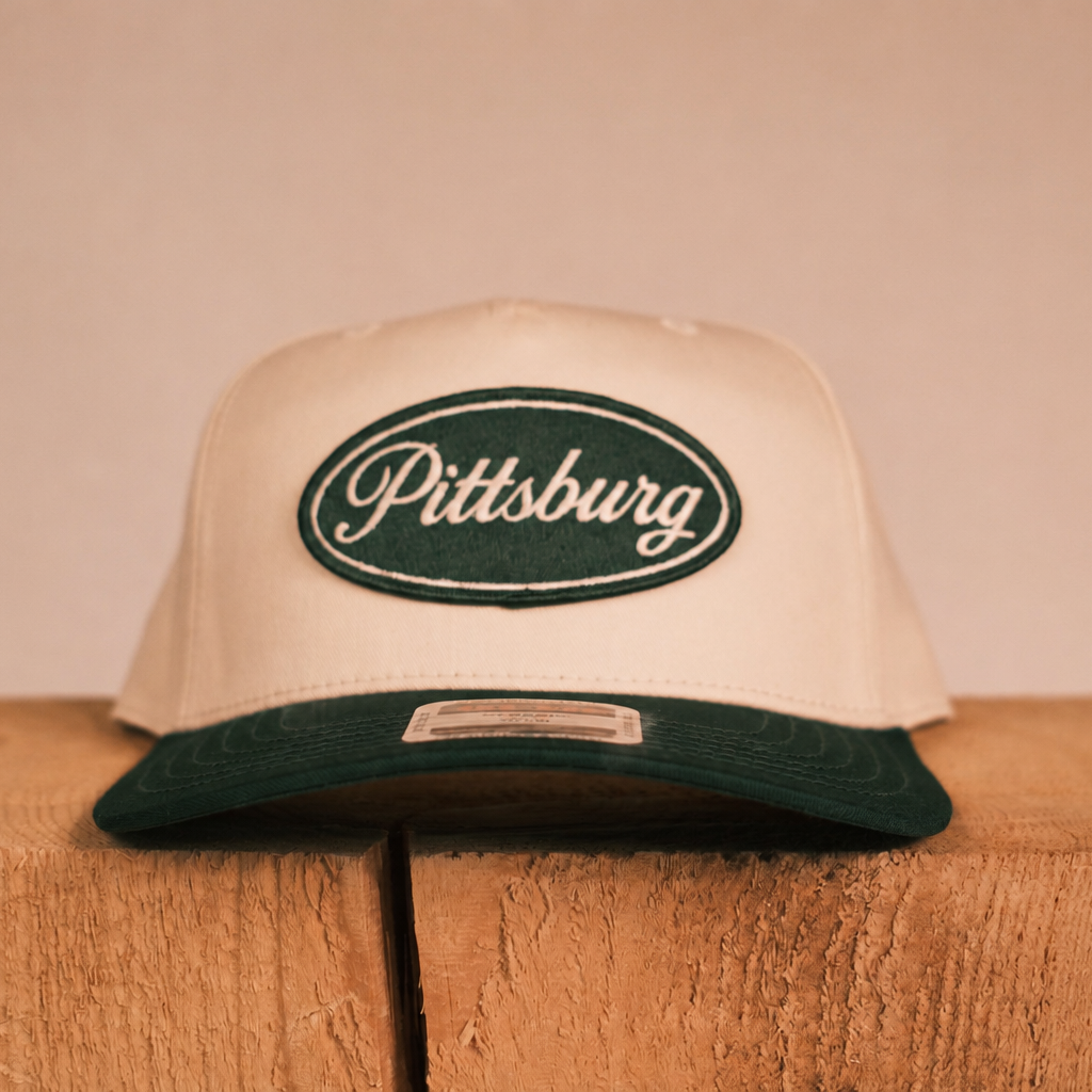 A cream-colored snapback cap with a dark green visor and a green embroidered oval patch on the front that reads 'Pittsburg' in white script. The cap is displayed on a rustic wooden block against a solid, warm beige background.