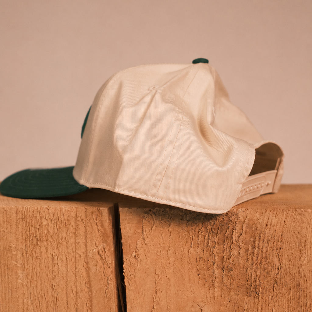 A cream-colored baseball cap with a dark green brim and matching green button, resting on a rustic wooden block against a solid beige background.