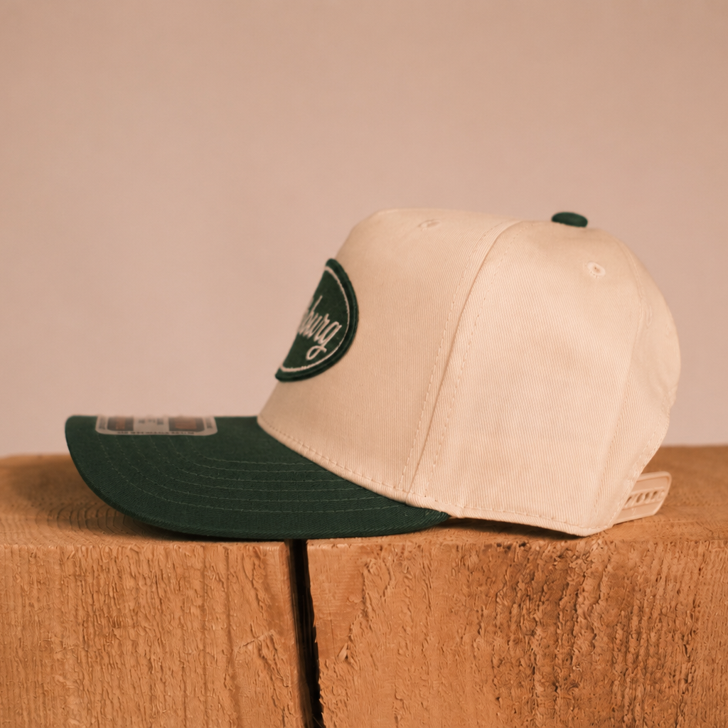 A cream-colored snapback cap with a dark green visor and a green circular patch on the side, displayed on a rustic wooden block against a solid beige background.