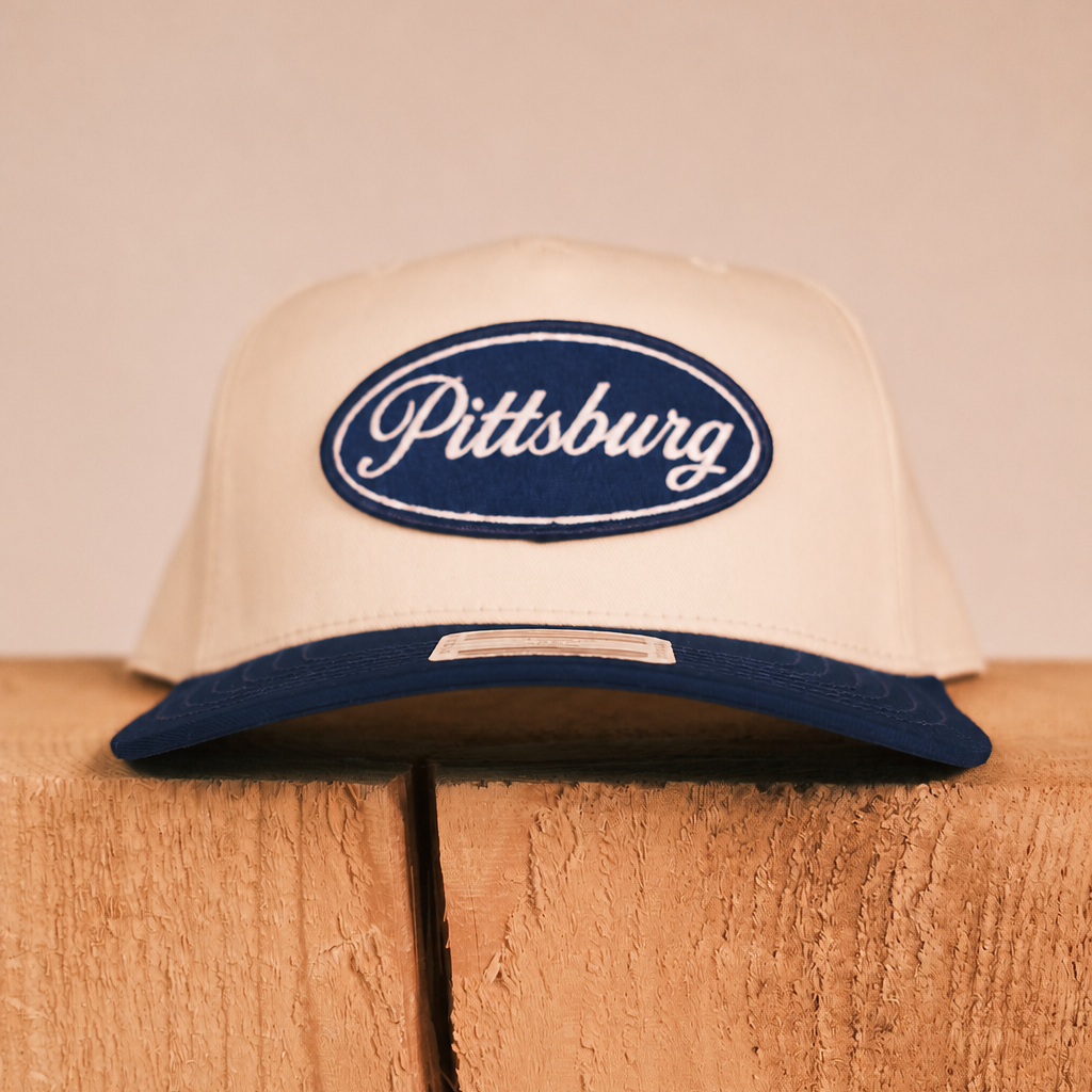 An off-white snapback cap with a navy blue brim and a navy blue oval patch on the front that reads 'Pittsburg' in white script, displayed on a rustic wooden block against a solid beige background.