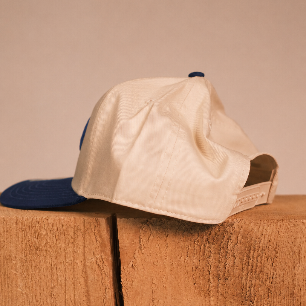 A cream-colored snapback cap with a dark blue brim, resting on a rustic wooden block against a neutral beige background.
