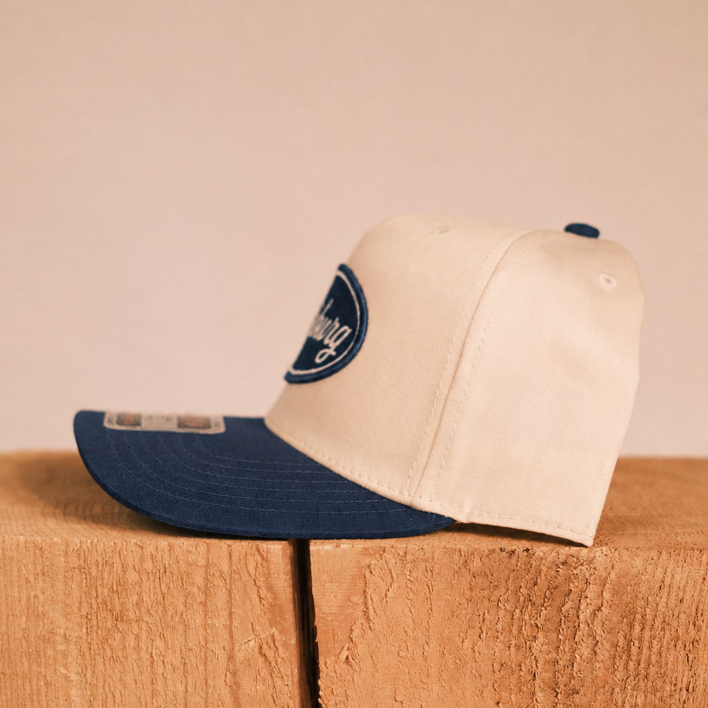 A two-tone baseball cap with a cream crown and a navy blue visor, resting on a wooden block against a solid beige background. The cap features a navy blue oval patch on the side with the text 'The Daily Grind'.
