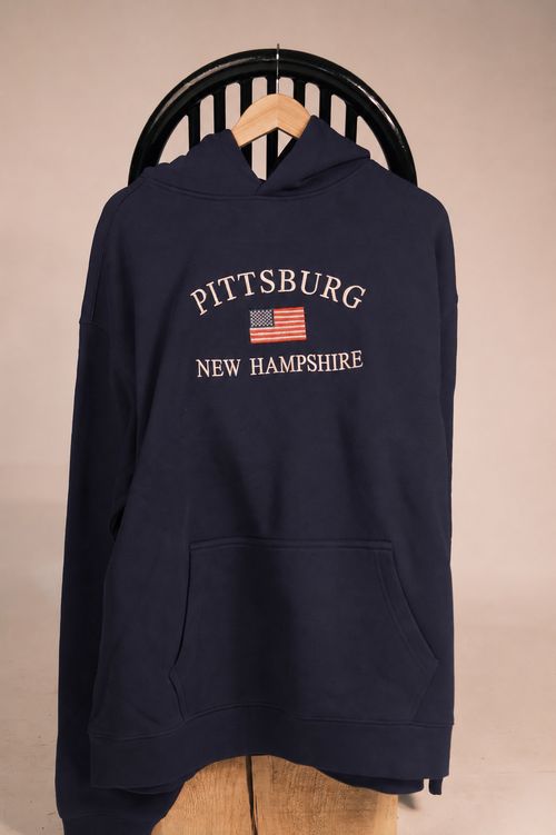A navy blue pullover hoodie hanging on a wooden hanger against a beige wall, featuring 'PITTSBURG NEW HAMPSHIRE' and a small American flag printed on the chest.