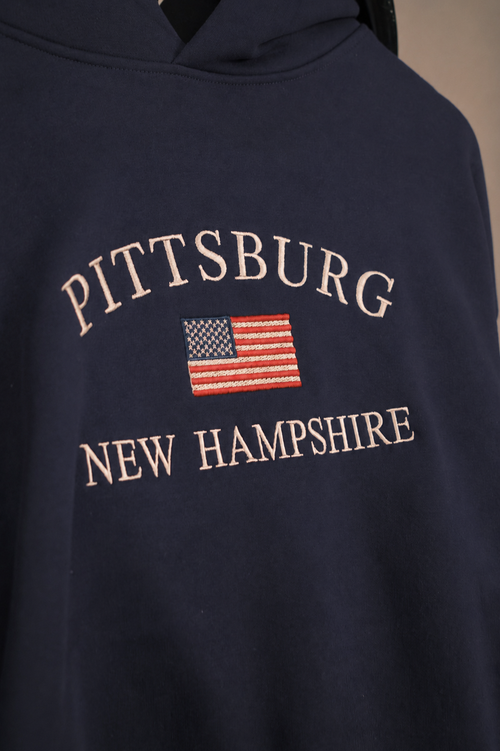 Close-up of a navy blue hoodie featuring 'PITTSBURG NEW HAMPSHIRE' in cream-colored arched embroidery and a small American flag graphic in the center.