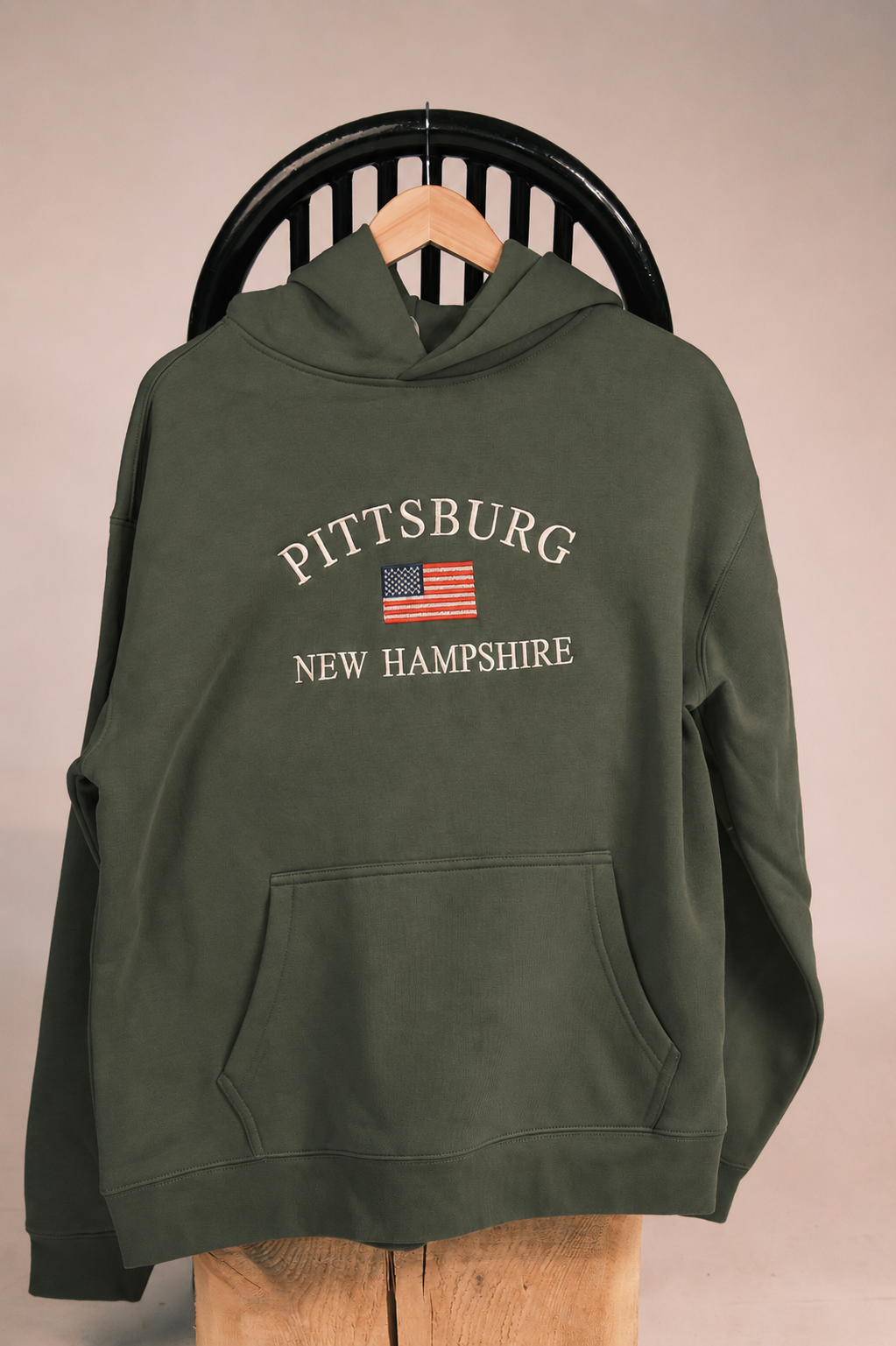 An olive green pullover hoodie with 'PITTSBURG NEW HAMPSHIRE' and an American flag graphic printed on the chest, hanging on a wooden hanger from a black chair.