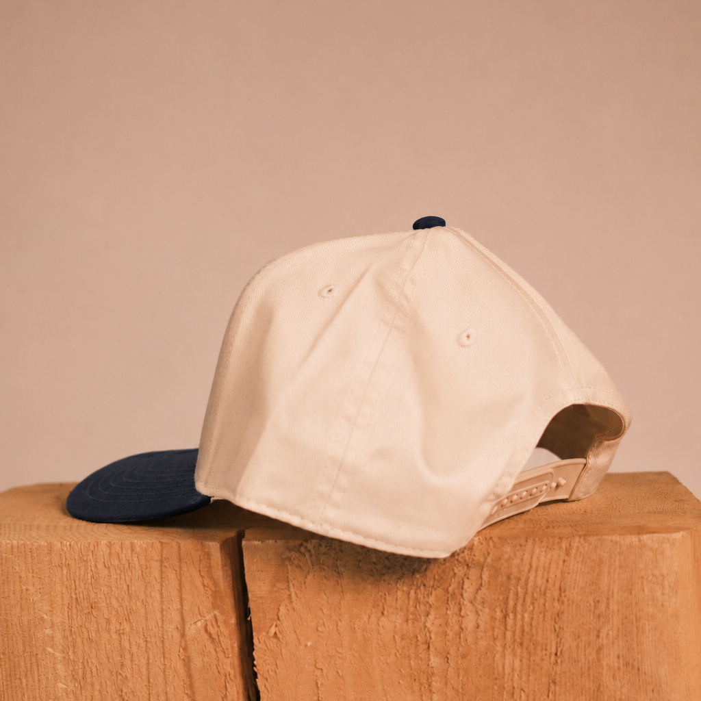 A cream-colored baseball cap with a dark navy blue brim, resting on a rustic wooden block against a solid beige background.
