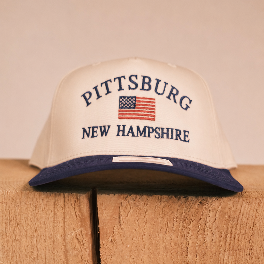 A cream-colored snapback cap with a navy blue brim, featuring 'PITTSBURG NEW HAMPSHIRE' and an American flag embroidered on the front, resting on a rustic wooden block against a neutral beige background.