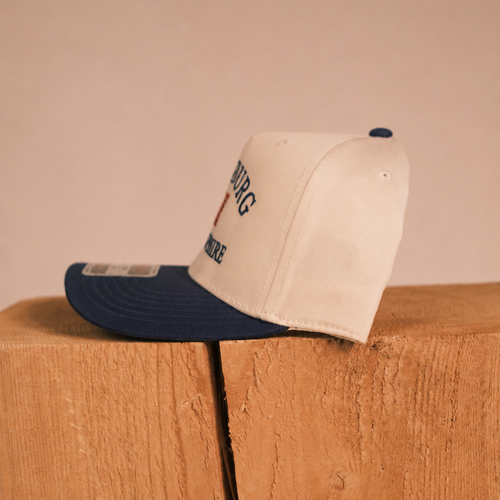 A cream-colored snapback cap with a navy blue visor and matching navy button, resting on a rustic wooden block against a solid beige background. The front of the cap features embroidered text reading 'BURG' and 'MUNICE' with a small red heart detail.