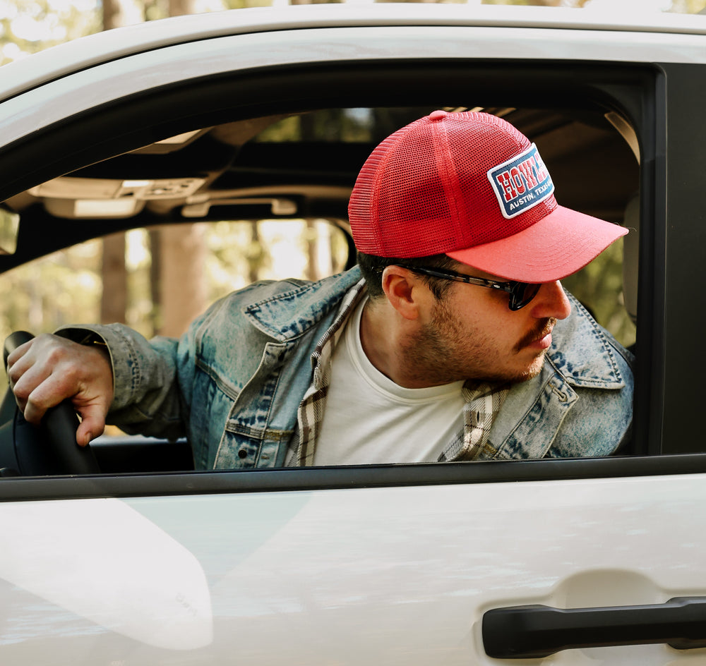 Man wearing a red cap with a logo, sitting inside a car with trees in the background