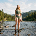 Woman in green swimsuit standing in a river with mountains and trees in the background