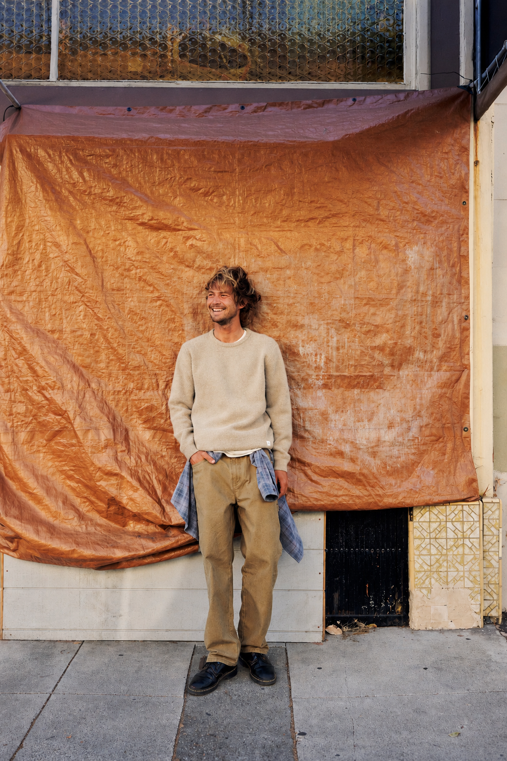 A man stands on a sidewalk in front of a large rust-orange tarp draped over a storefront, wearing a beige sweater, tan pants, and dark shoes, with a blue denim jacket hanging from his waist.