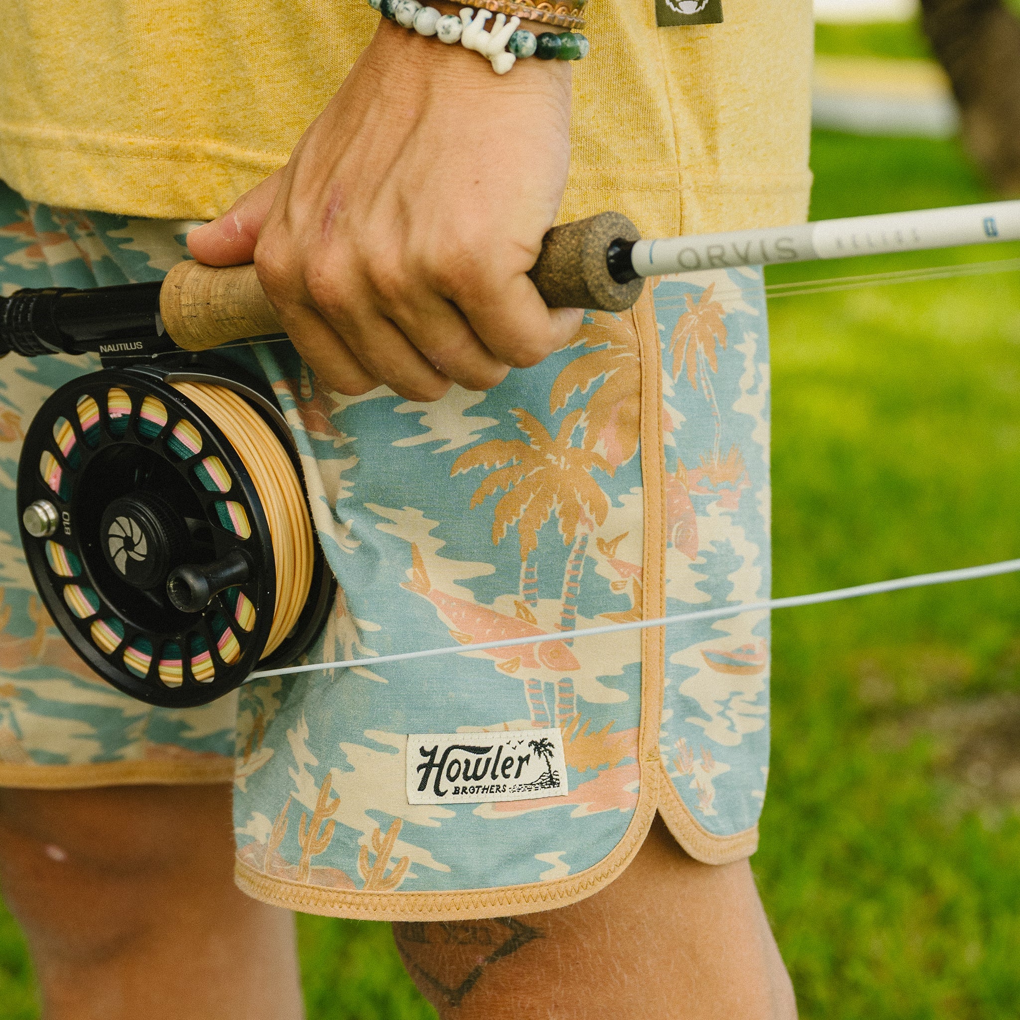 Person holding a fly fishing reel with 'Howler' branding on shorts, outdoors.