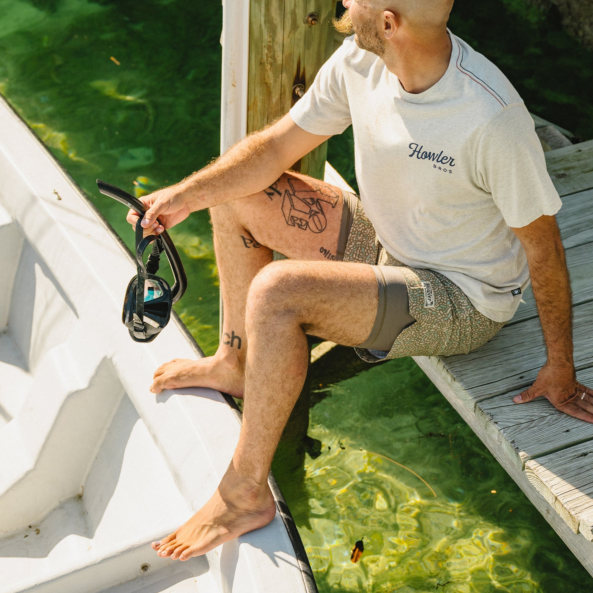 Man sitting on a dock by a body of water, wearing a Howler Bros t-shirt.