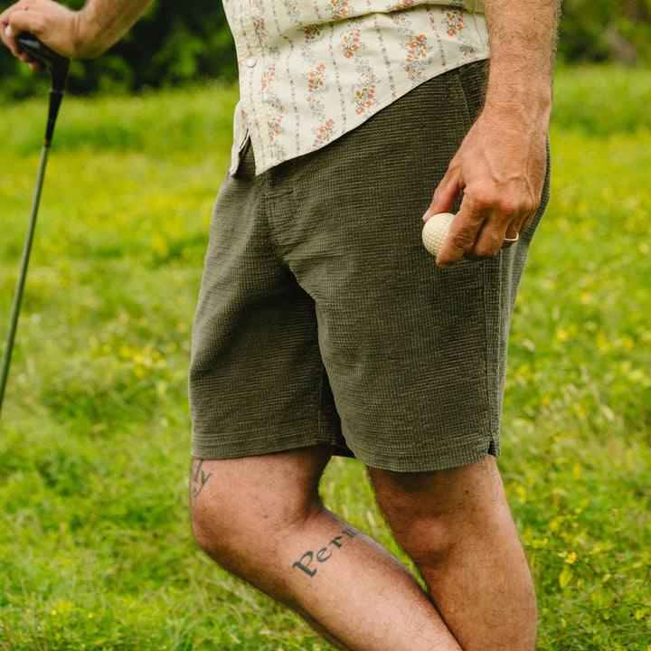 Person wearing green shorts holding a golf club and ball on a grassy field