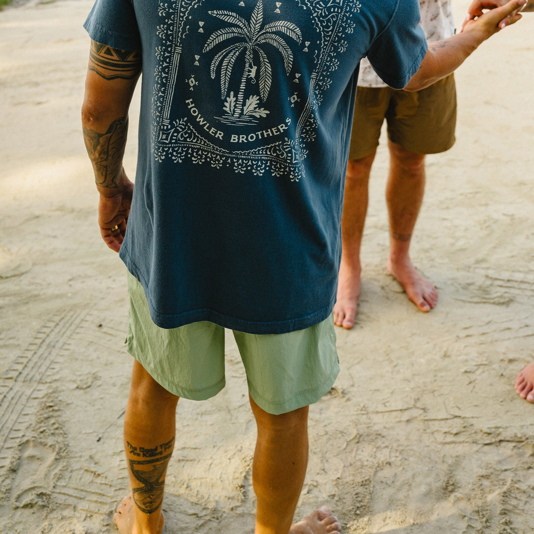 Person wearing a blue t-shirt with a palm tree design on a sandy beach.