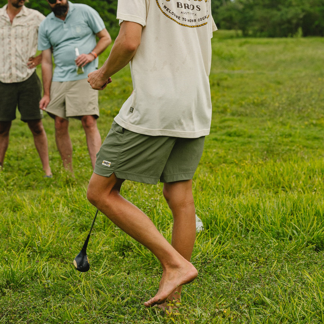 Person walking barefoot on grass with a golf club, wearing a 'Bros' t-shirt and green shorts.