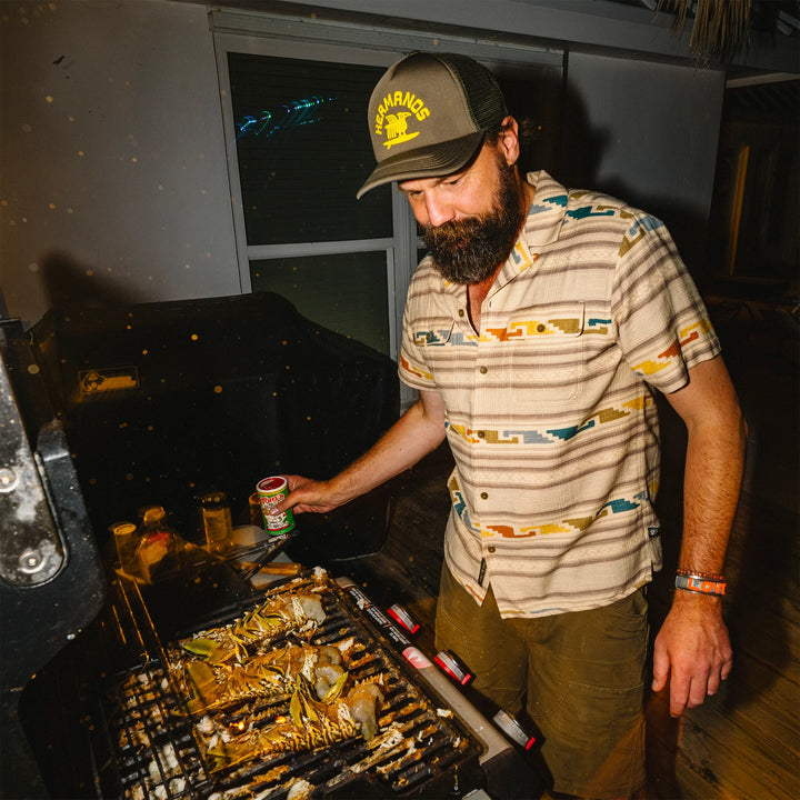 Man grilling outdoors at night, wearing a cap and patterned shirt.