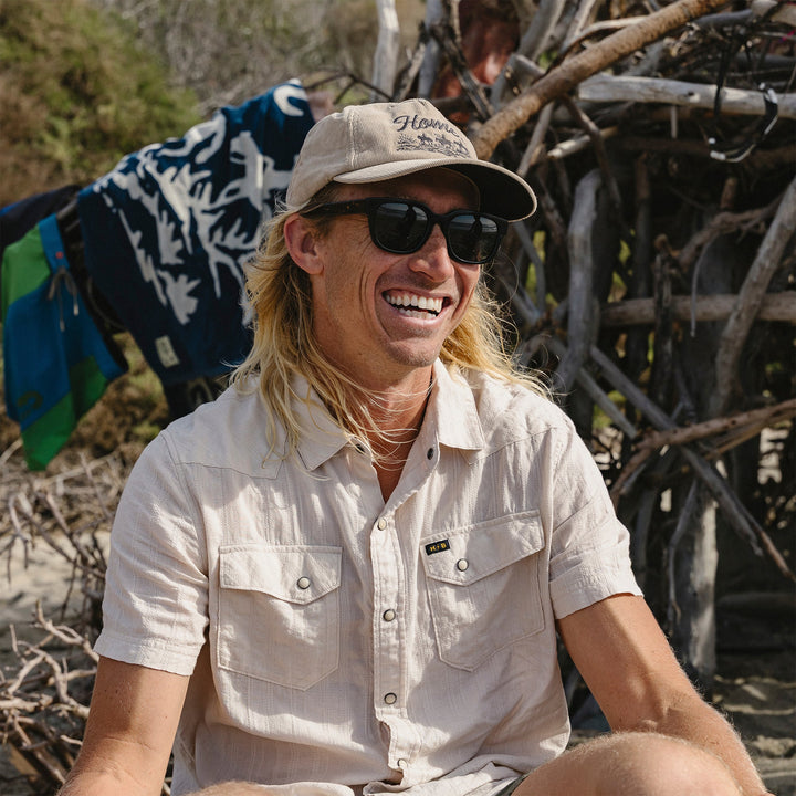Man wearing sunglasses and a cap sitting outdoors with a rustic background