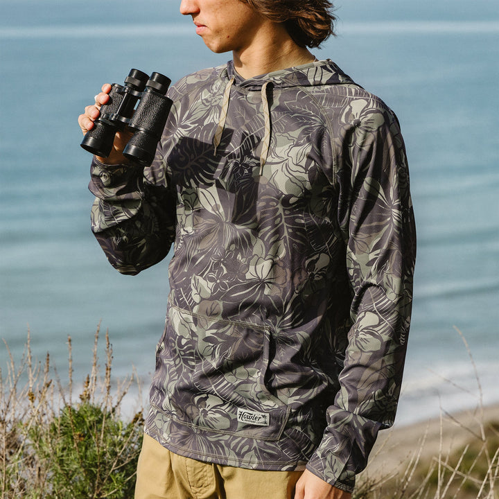 Person wearing a floral-patterned jacket holding binoculars by the ocean.