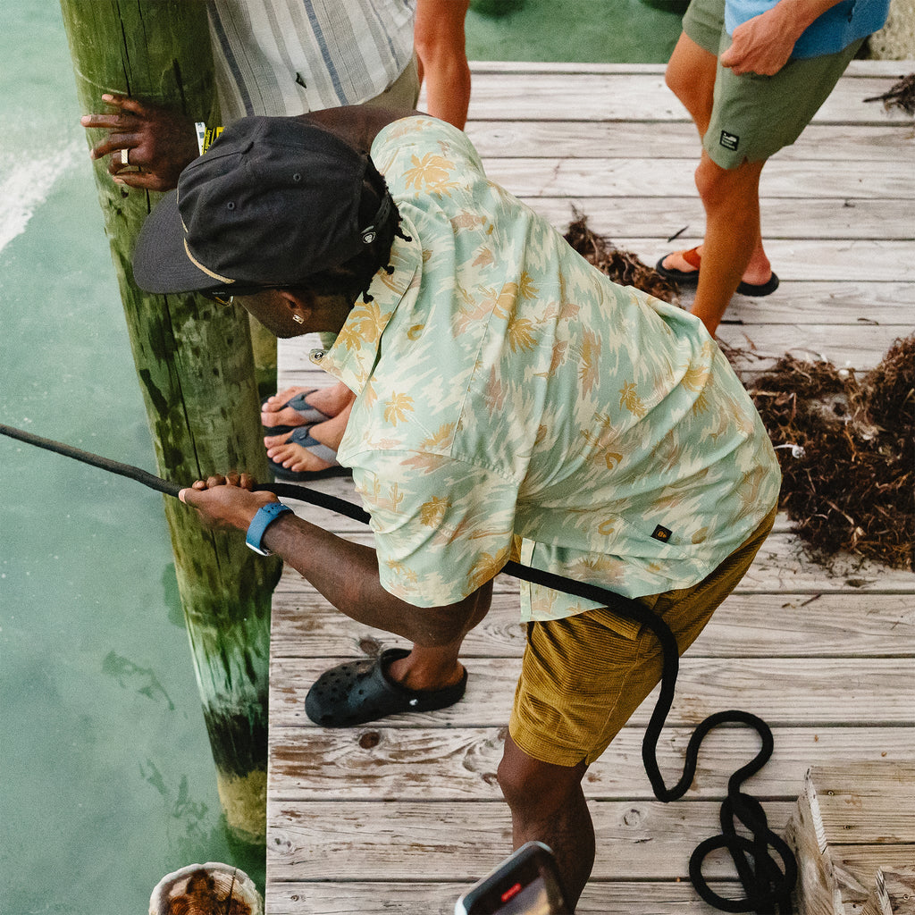 Person on a dock tying a rope to a post, wearing a floral shirt and yellow shorts.