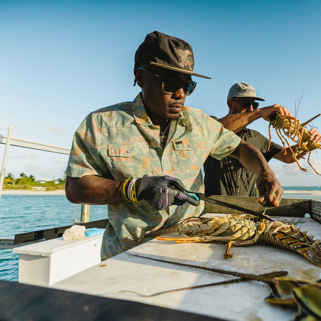 Two men on a boat, one cutting a crab with shears, with water and sky in the background.