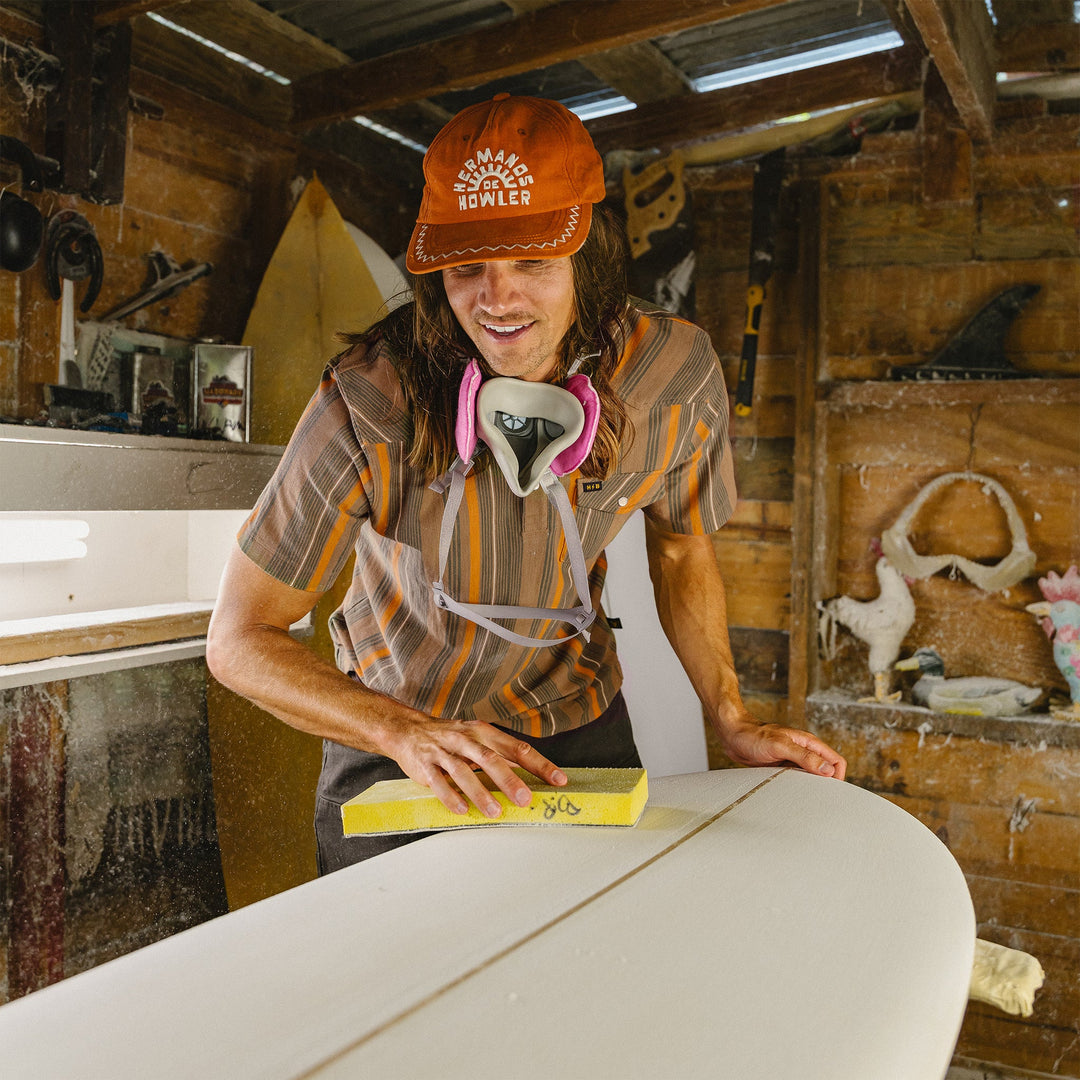 Person sanding a surfboard inside a workshop with tools and equipment visible.