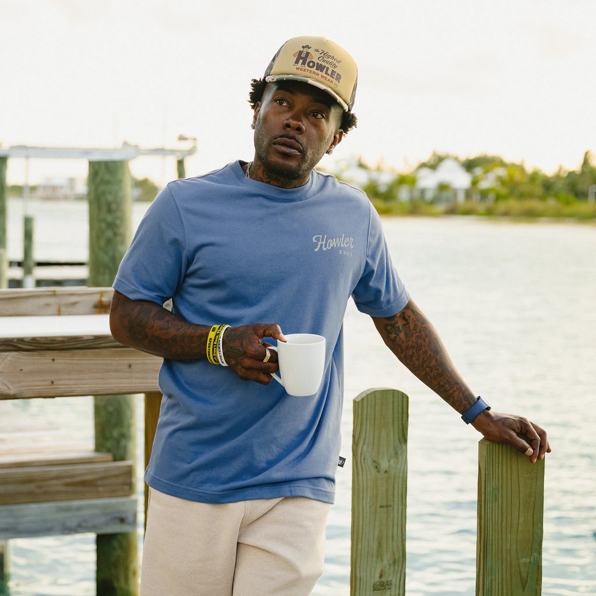 Man wearing a blue shirt and beige cap holding a white mug by a waterfront.