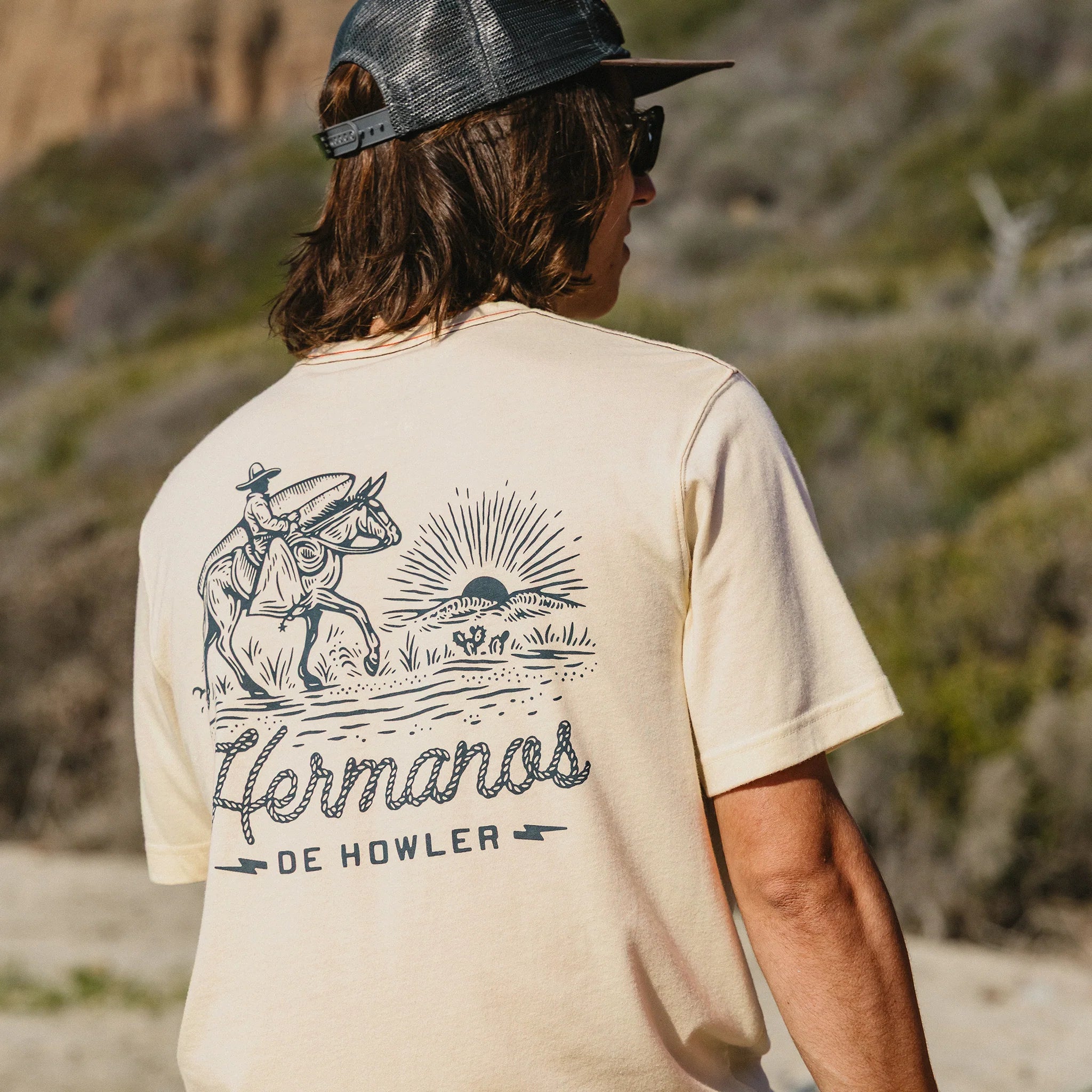 Person wearing a beige t-shirt with a graphic design and text, standing in a natural setting.