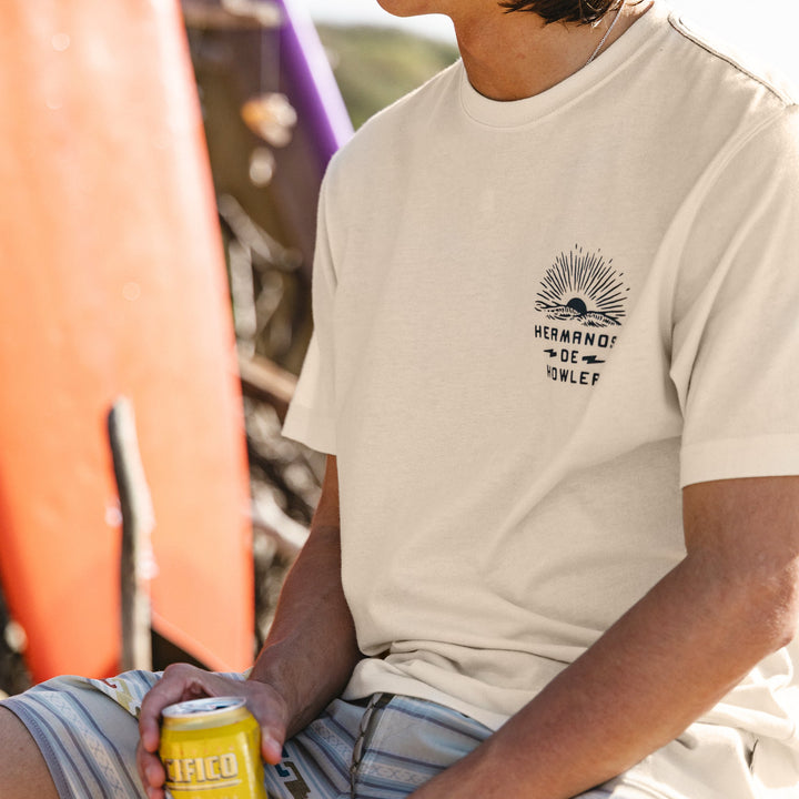 Person wearing a beige t-shirt with a logo, holding a can, with a blurred outdoor background.