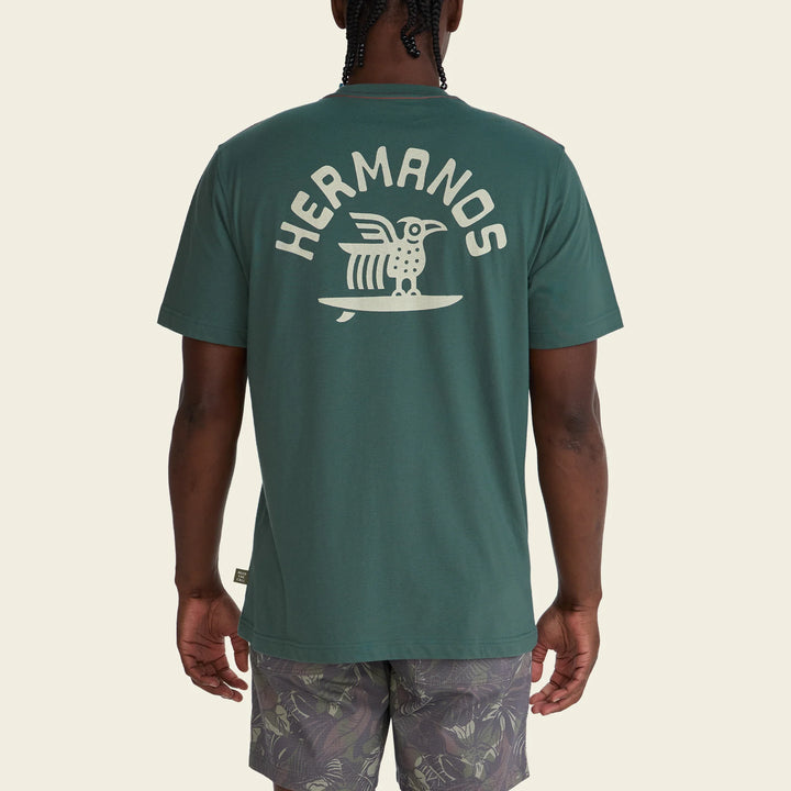 Person wearing a green t-shirt with 'Hermanos' logo on the back, standing against a beige background